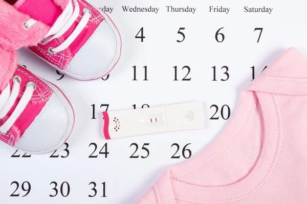 Tes kehamilan dengan hasil positif dan pakaian untuk bayi yang baru lahir pada kalender, mengharapkan bayi — Stok Foto