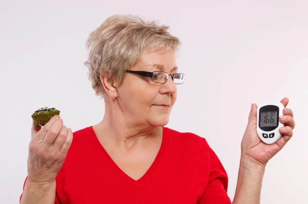 Surprised senior woman holding glucometer and fresh cupcake, measuring and checking sugar level, concept of diabetes — Stockfoto