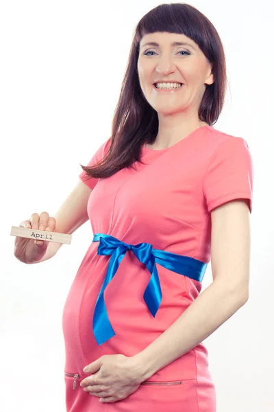 快乐的孕妇穿着蓝色缎带的粉色连衣裙 在木制立方体上写着 April 对新生儿的期望和家庭的扩大 — 图库照片