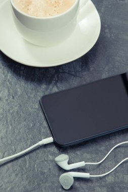 Bağlı kulaklıklı siyah akıllı telefon ve sütlü kahve. Müzikle gevşeme zamanı