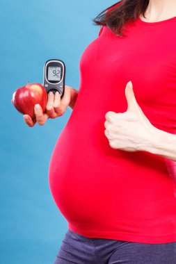 Hamile kadın baş parmağını kaldırıyor, taze elma ve glikoz ölçer tutuyor ve şeker seviyesini ölçüyor. Hamilelik süresince diyabet kavramı, yeni doğmuş bir bebek bekliyor.