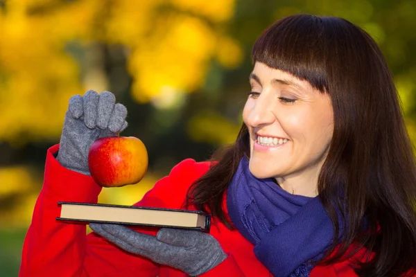 Happy woman holding fresh apple and book in autumnal park, autumn concept — Stockfoto