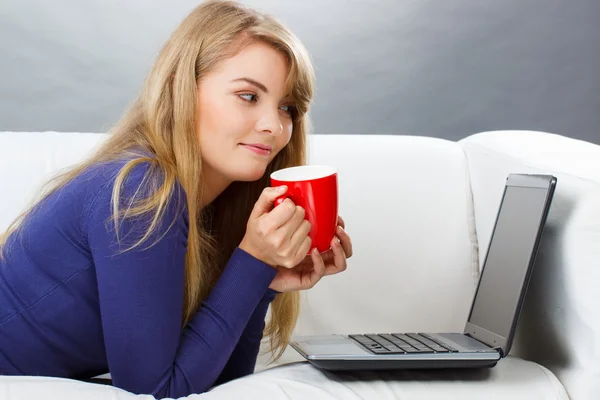 Счастливая женщина с чашкой чая, используя ноутбук, лежащий на диване, современные технологии — стоковое фото