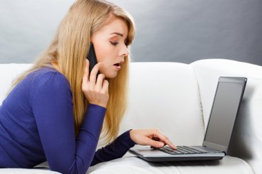 Woman talking on mobile phone and using laptop lying on sofa, modern technology