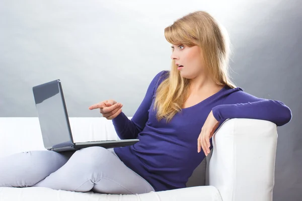 Mujer sorprendida sentada en el sofá y mostrando el ordenador portátil, la tecnología moderna — Foto de Stock