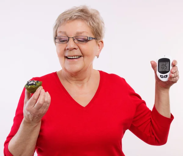 Счастливая пожилая женщина держит глюкометр и свежий кекс, измерения и проверки уровня сахара, концепция диабета — стоковое фото