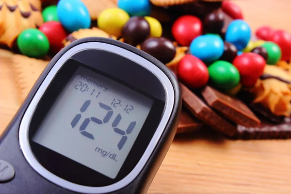 Medidor de glucosa con un montón de dulces en la superficie de madera, diabetes y alimentos poco saludables — Foto de Stock