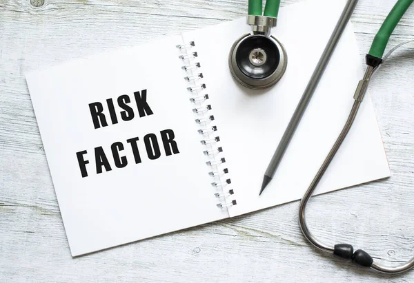 RISK FACTORS is written in a notebook on a light wooden table next to pencil and a stethoscope. Medical concept