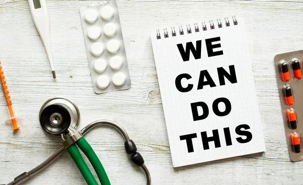 WE CAN DO THIS is written in a notebook on a white table next to pills and a stethoscope. Medical concept