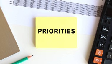 Laptopun yanındaki sarı yapışkan notlardaki yazı ve beyaz ofis masasındaki hesap makinesi..