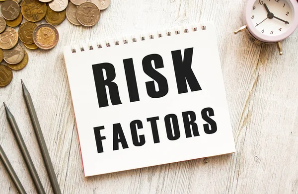 RISK FACTORS text on a sheet of notepad. Coins are scattered, pencils on a gray wooden background. Financial concept.