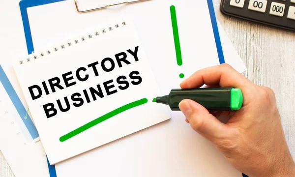 A hand with a marker writes the text DIRECTORY BUSINESS in a notebook on the desktop. View from above. Business concept.