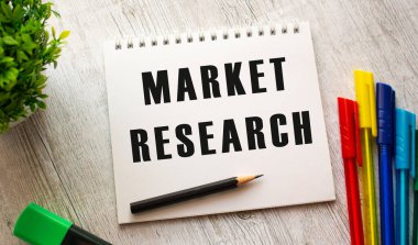 Bir yay üzerinde beyaz bir çarşaf üzerinde MARKET RESEARCH yazılı bir defter renkli kalemlerle ahşap bir masada yatıyor. İş konsepti.
