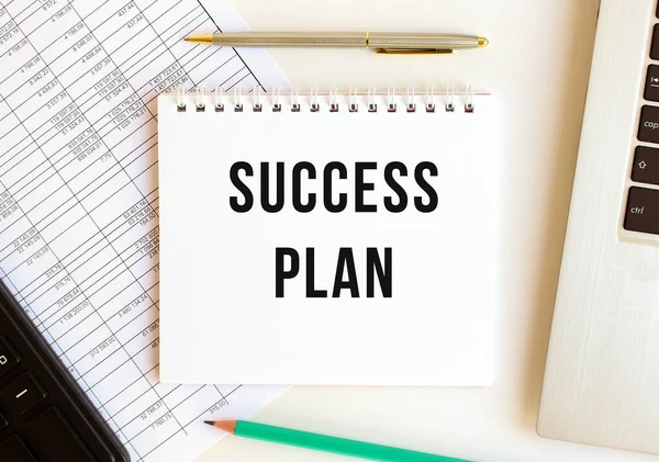 Bilgisayarın, hesap makinesinin ve ofis malzemelerinin yanında beyaz bir arkaplanda SUCCESS PLAN yazılı not defteri. İş konsepti.