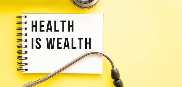 Text HEALTH IS WEALTH on notebook with stethoscope on yellow background. Medical concept.