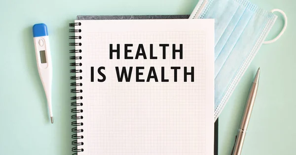 Notepad, medical mask, thermometer and pen on a blue background. HEALTH IS WEALTH text in a notebook. Medical concept.