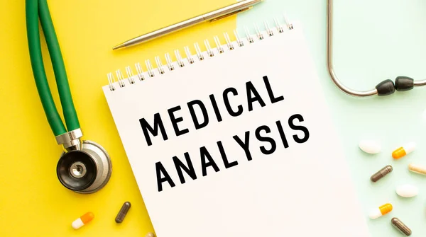 MEDICAL ANALYSIS is written in a notebook on a color table next to pills and a stethoscope. Medical concept