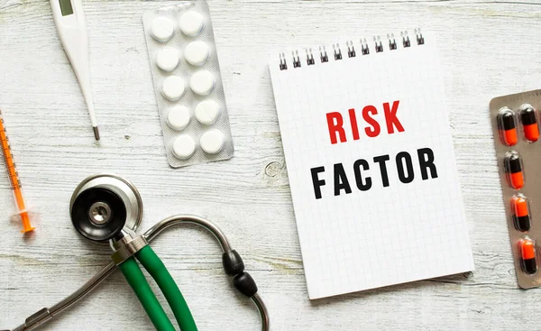 RISK FACTOR is written in a notebook on a white table next to pills and a stethoscope. Medical concept
