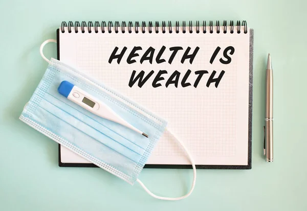 A medical protective mask and a thermometer are on the notepad. HEALTH IS WEALTH text in a notebook. Medical concept.