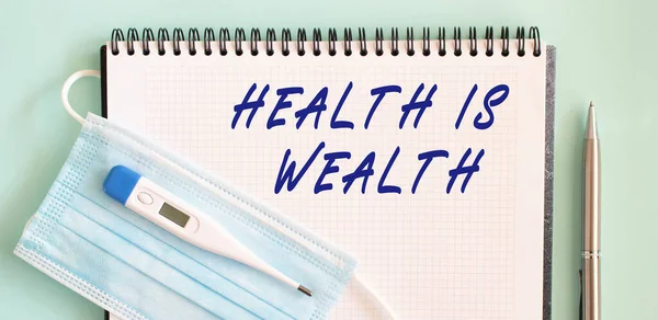 A medical protective mask and a thermometer are on the notebook. HEALTH IS WEALTH text in a notebook. Medical concept.