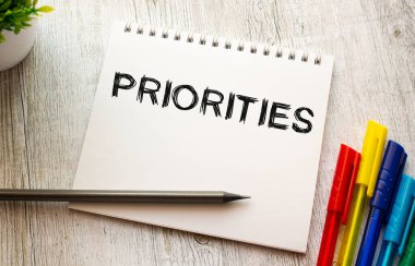 Bir yay üzerinde beyaz bir çarşaf üzerinde PRIORITIES yazılı bir defter renkli kalemlerle ahşap bir masanın üzerinde yatar. İş konsepti.