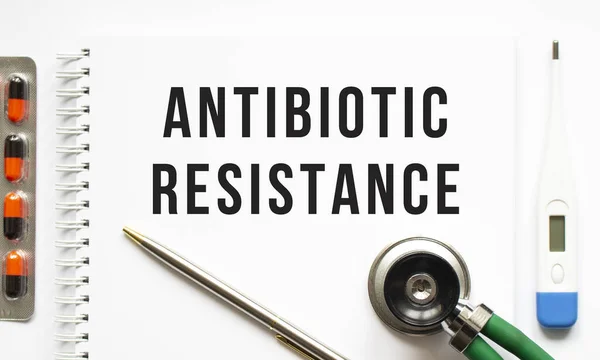 ANTIBIOTIC RESISTANCE is written in a notebook on a white table next to pills and a stethoscope. Medical concept