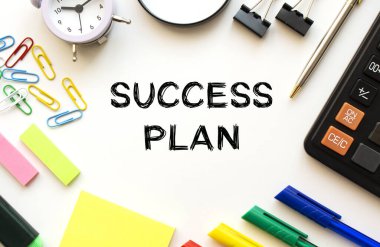 Hesap makinesi, büyüteç, renkli kalemler ve diğer kağıtlarla dolu beyaz ofis masası. SUCCESS Planı üzerine metin. Yukarıdan bak. İş konsepti.