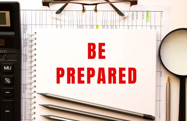 Office desk with financial papers, magnifying glass, calculator, glasses. Notepad page with the text BE PREPARED. View from above. Business concept.