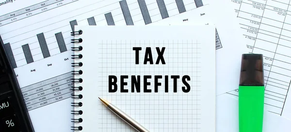 Text TAX BENEFITS on the page of a notepad lying on financial charts on the office desk.