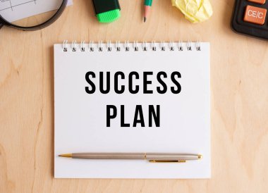 Hesap makinesi ve ofis malzemelerinin yanında, ahşap bir masanın üzerinde SUCCESS PLAN yazılı not defteri. İş konsepti.