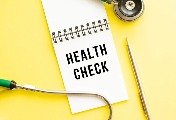 HEALTH CHECK is written in a notebook on a color table next to pen and a stethoscope. Medical concept