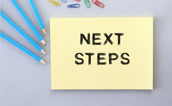 NEXT STEPS text in notebook on gray background next to pencils and paper clips. Concept.