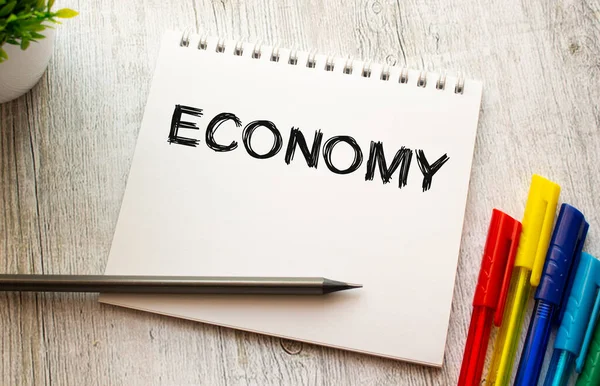 Bir yay üzerinde beyaz bir çarşaf üzerinde ECONOmy yazılı bir defter renkli kalemlerle ahşap bir masada yatıyor. İş konsepti.