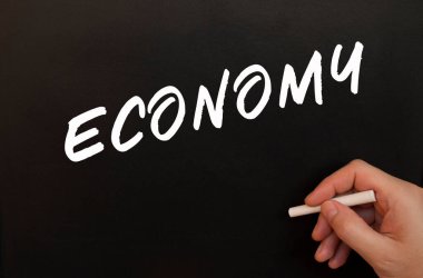 Erkek el tebeşirle ECONOmy kelimelerini kara tahtaya yazar. İş konsepti.