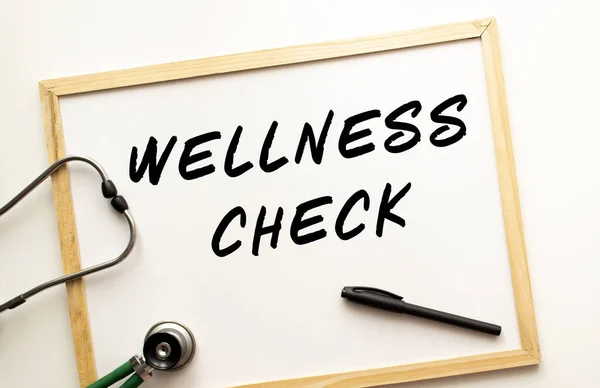 The text WELLNESS CHECK is written on a white office board with a marker. Nearby is a stethoscope. Medical concept.