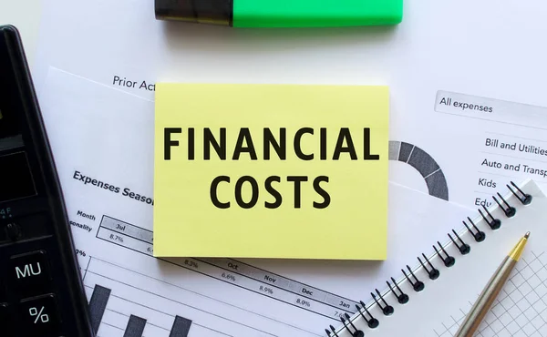 Text FINANCIAL COSTS on the page of a notepad lying on financial charts on the office desk. Near the calculator. Business concept.