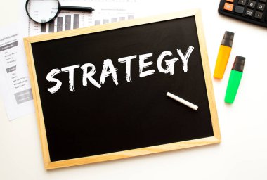 Bir tahtaya tebeşirle yazılmış STRATEGY metni. Ofis masası ve ofis malzemeleri. İş konsepti.