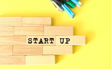Ahşap bloklar sarı arka planda kalem ve kalemlerin yanına istiflenmiş. Tahta bir blokta STARTUP metni. İş konsepti