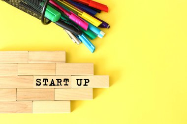 Ahşap bloklar sarı arka planda kalem ve kalemlerin yanına istiflenmiş. Tahta bir blokta STARTUP metni. İş konsepti
