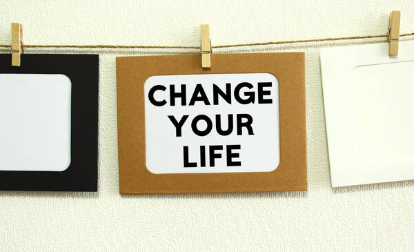 Крафт бумажная рамка висит на шнуровке на белом фоне стены с пространством. В рамке написан текст Изменить свою жизнь.