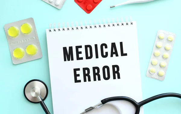 The text MEDICAL ERROR is written in a notebook that lies next to the stethoscope, medicine on a blue background. Medical concept