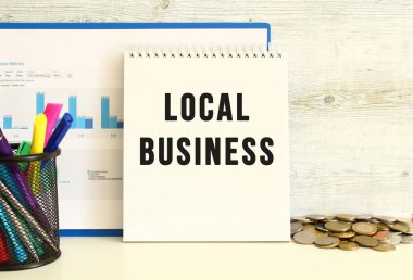 Üzerinde LOCAL BUSINESS yazan bir defter, mali tabloların olduğu bir klasörle birlikte duvara yaslanıyor. Kırtasiye ve madeni paraların yanında. İş konsepti.