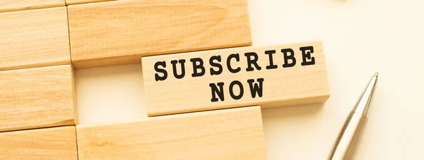 SUBSCRIBE NOW text on a strip of wood lying on a white table with a metal pen. Concept.