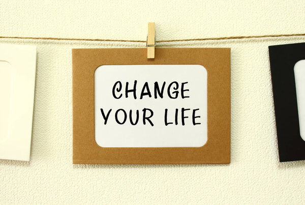 Крафт бумажная рамка висит на шнуровке на белом фоне стены с пространством. В рамке написан текст Изменить свою жизнь.