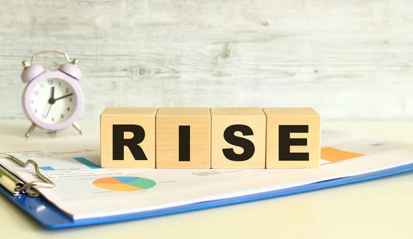 Wooden cubes lie on a folder with financial charts on a gray background. The cubes make up the word RISE. — Stock Photo, Image