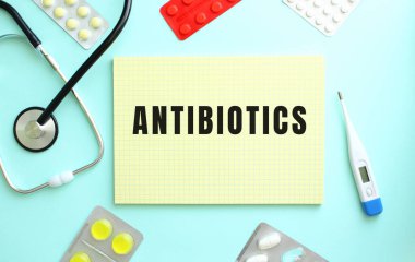 ANTIBIOTICS metni, steteskopun yanında duran sarı bir deftere yazılmıştır, mavi arka planda bulunan bir ilaç. Tıbbi konsept