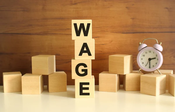 Four wooden cubes stacked vertically on a brown background form the word WAGE. Cubes are scattered nearby and there is a clock. Front View Concepts