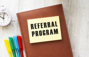 Bilgisayarın, hesap makinesinin ve ofis malzemelerinin yanında beyaz bir arkaplanda REFERRAL PROGRAM metni olan not defteri. İş konsepti.