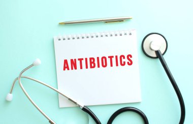 Kırmızı metinli beyaz not defteri ANTIBIOTICS ve mavi arka planda steteskop. Tıbbi konsept