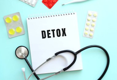 DETOX metni steteskopun yanında duran beyaz bir not defterine ve mavi arka planda bulunan haplara yazılmıştır..